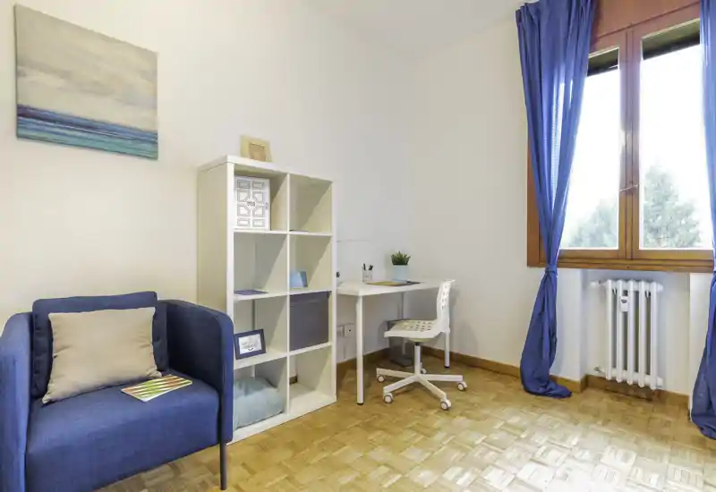 Bright, neatly staged living area with a blue armchair, shelving unit and a small workspace by the window — ideal for relaxing or working from home.