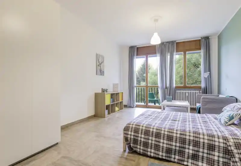 Camera da letto luminosa e arredata con gusto: letto matrimoniale con copriletto a quadri, piccolo angolo conversazione vicino alla finestra e luce naturale da un balcone.