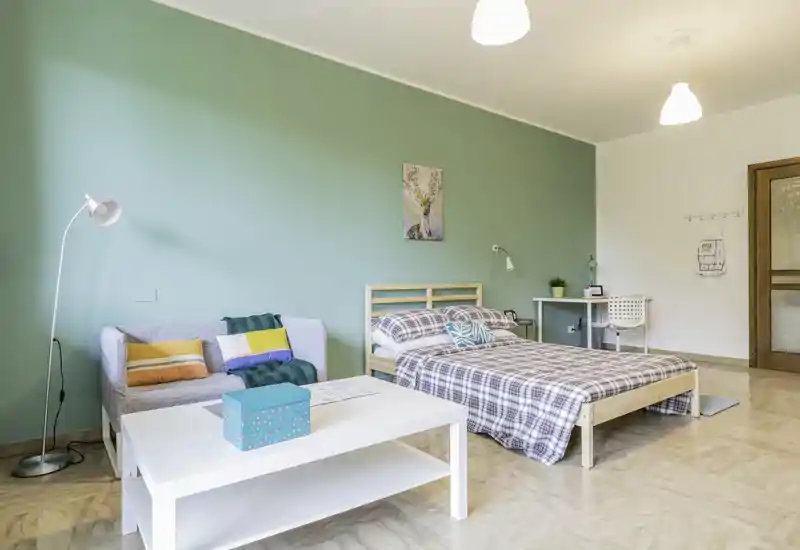 Camera da letto luminosa e ben allestita con letto in legno, biancheria a quadri accogliente, un piccolo divano e tavolino — parete verde chiaro e illuminazione moderna donano atmosfera calda.