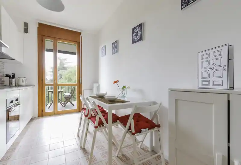 Bright, compact kitchen with a small dining table set for two, red seat cushions, modern white cabinets and a glass door opening to a small balcony — ideal for cozy breakfasts.