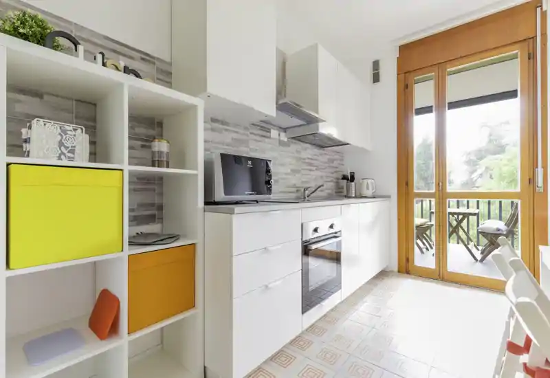 Bright modern kitchenette with white cabinets, built-in oven and a large glass door opening to a small balcony with seating — tidy storage and colorful accents create a cheerful feel.