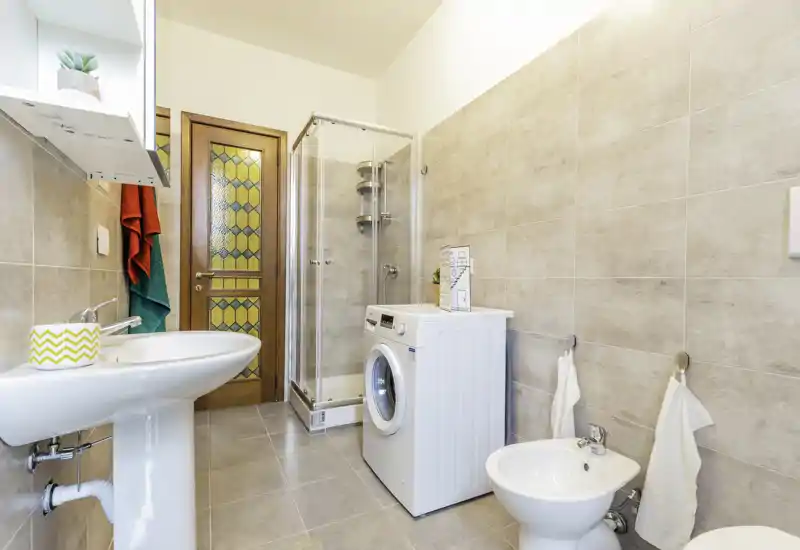 Bright, modern bathroom with a shower cabin, washing machine, sink and bidet. Neutral tiles and good lighting create a clean, functional space.