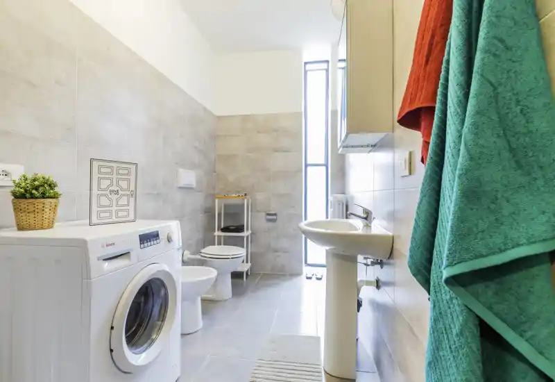 Bright, modern bathroom with a pedestal sink, toilet, bidet and a front-loading washing machine; light tiles and a tall frosted window provide abundant natural light.
