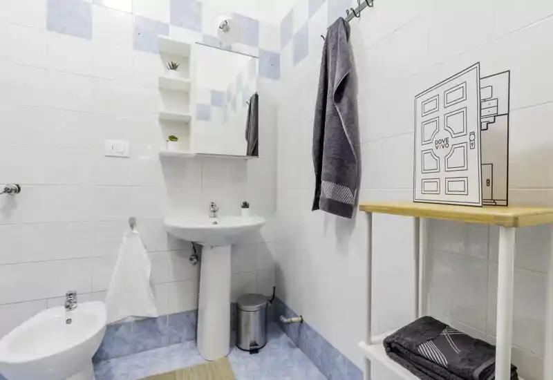 Clean, bright bathroom with pedestal sink, bidet and tiled walls in soft blue and white tones. Towels hang neatly and a small shelving unit provides storage.