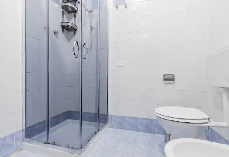 Clean, bright bathroom with a glass-enclosed shower, wall-mounted toilet and light blue tiled floor and lower walls.