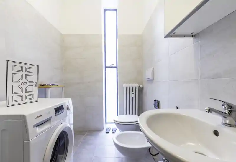 Bright modern bathroom with a sink, bidet, toilet and a washing machine, flooded with natural light from a tall narrow window.