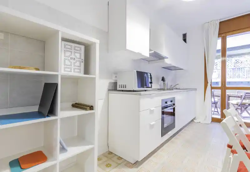 Bright, modern kitchenette with white cabinets, integrated oven and a small countertop appliance. Natural light from the balcony door enhances the clean, airy atmosphere.