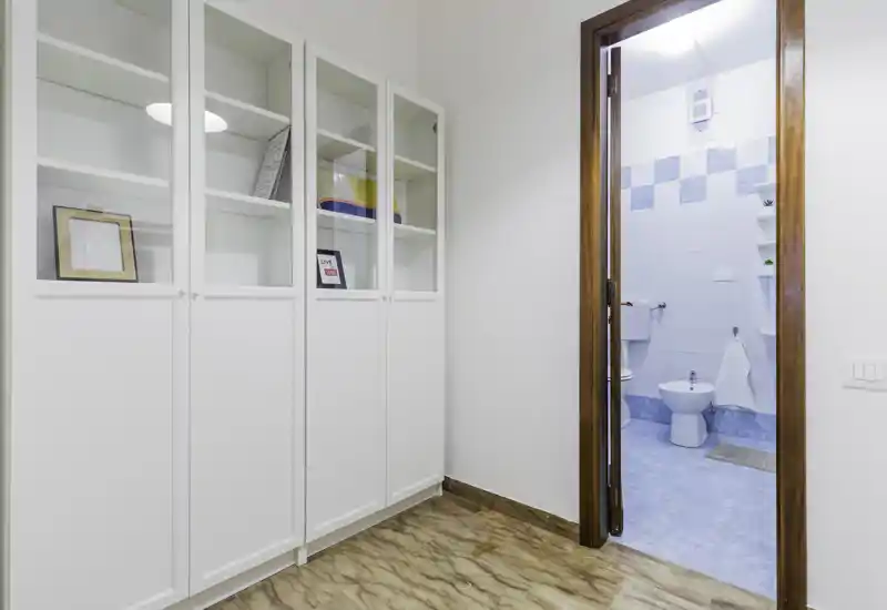 Bright, tidy entrance or small corridor with built-in white storage cabinets and a view into a tiled bathroom. Polished floor and clean minimalist styling make this space welcoming.