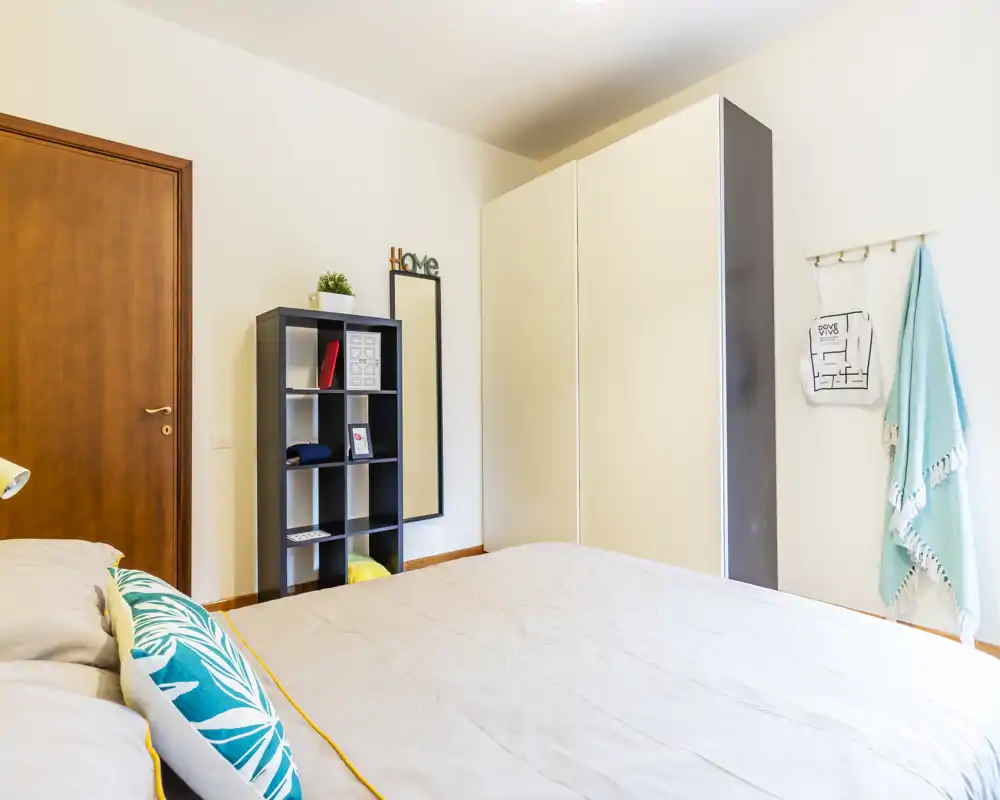 Camera da letto luminosa e moderna con un ampio letto in primo piano, cuscini decorativi e un armadio minimalista con scaffalatura — ideale per annunci in affitto che valorizzano l’area notte.