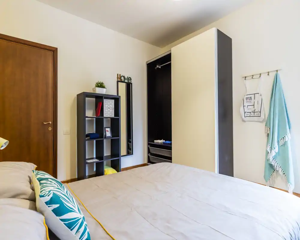 Camera da letto luminosa e ordinata con un grande letto in primo piano, cuscini decorativi e un armadio aperto con scaffalatura sullo sfondo.