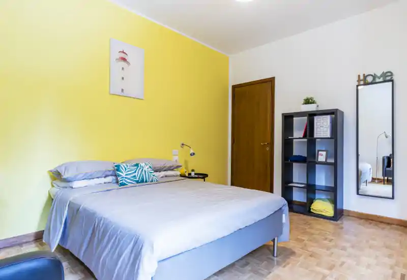 Camera da letto luminosa e ben arredata con un letto matrimoniale davanti a una parete gialla, cuscini decorativi, una libreria e uno specchio a figura intera. L'ambiente è accogliente e moderno, con buona illuminazione naturale.