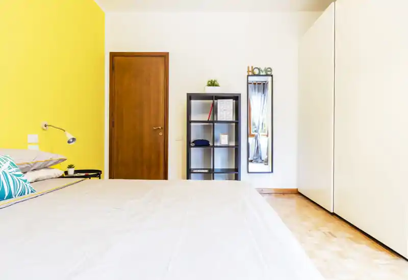 Camera da letto luminosa e moderna con ampio letto in primo piano, parete gialla a contrasto, lampada da comodino e una semplice libreria. Spazio pulito e ben illuminato con pavimento in parquet e armadio a tutta altezza.