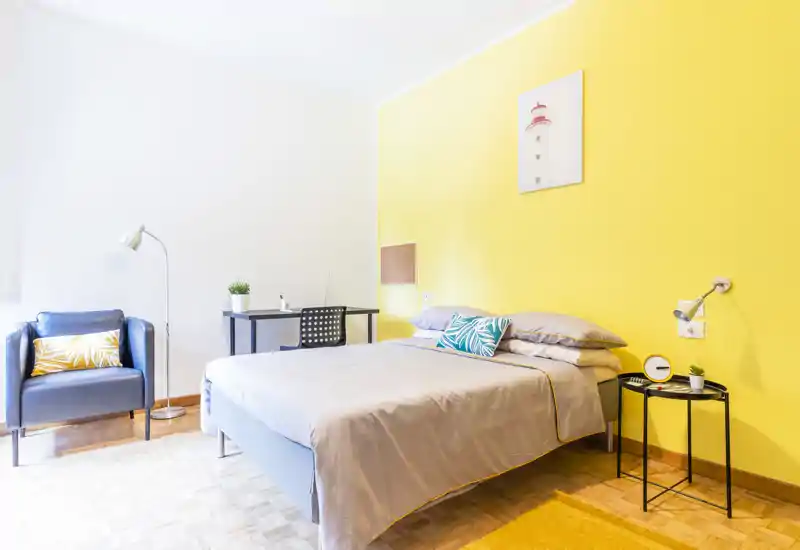 Camera da letto moderna e luminosa con letto matrimoniale, parete colorata e tessili accoglienti. Una scrivania e una poltrona creano un angolo pratico per lavoro o relax.