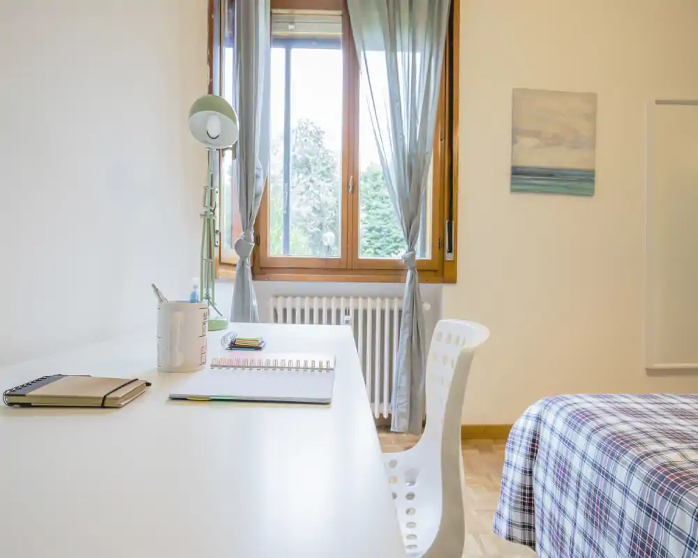 Rincón de dormitorio luminoso y ordenado con escritorio junto a una gran ventana y una cama con colcha a cuadros. La luz natural y la decoración sencilla crean un espacio tranquilo para dormir y trabajar.