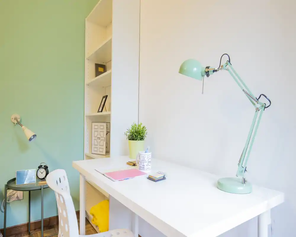 Rincón de dormitorio moderno y luminoso con escritorio blanco, lámpara ajustable verde menta, pequeña planta y estantería — perfecto como zona de estudio en la habitación.