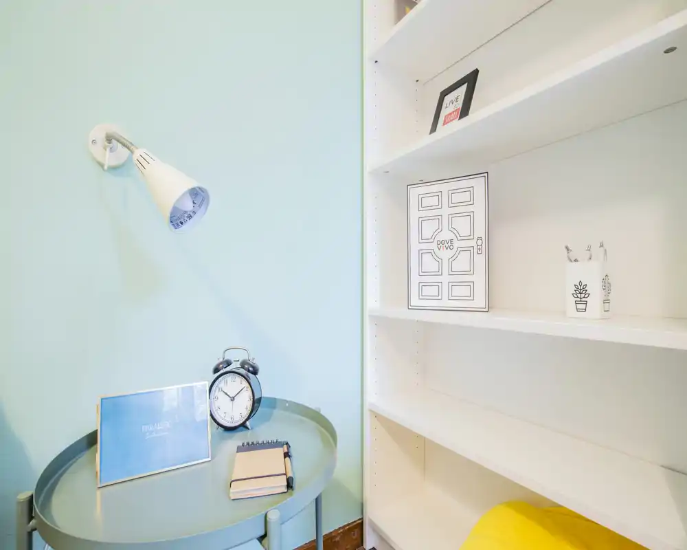 Rincón de dormitorio luminoso y de estilo sencillo con mesita, despertador y lámpara de pared; estanterías y elementos decorativos crean un ambiente moderno y tranquilo.