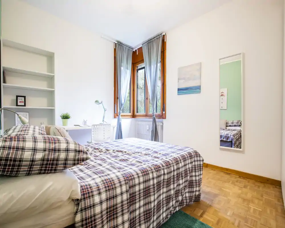 Dormitorio luminoso y ordenado con una cama doble vestida con ropa de cuadros, un escritorio junto a la ventana y suave luz natural de las ventanas con marco de madera.