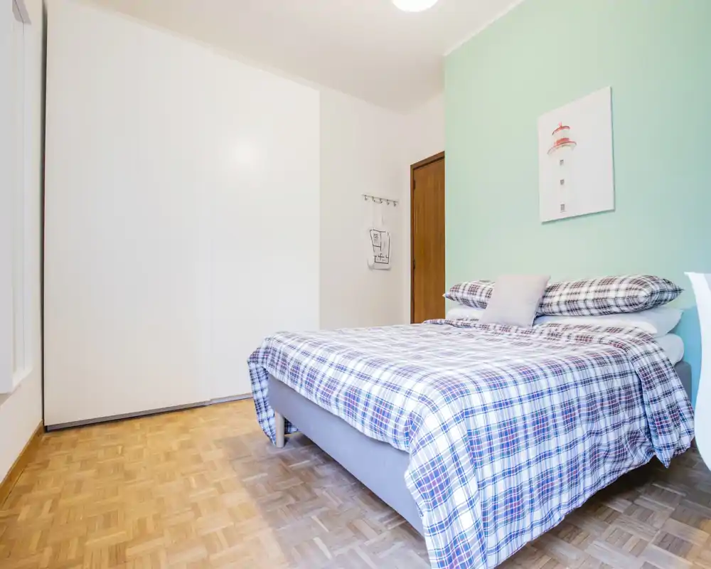 Dormitorio luminoso y ordenado con cama doble vestida con ropa de cuadros, cojines suaves y una pared verde menta que aporta un ambiente fresco y acogedor.