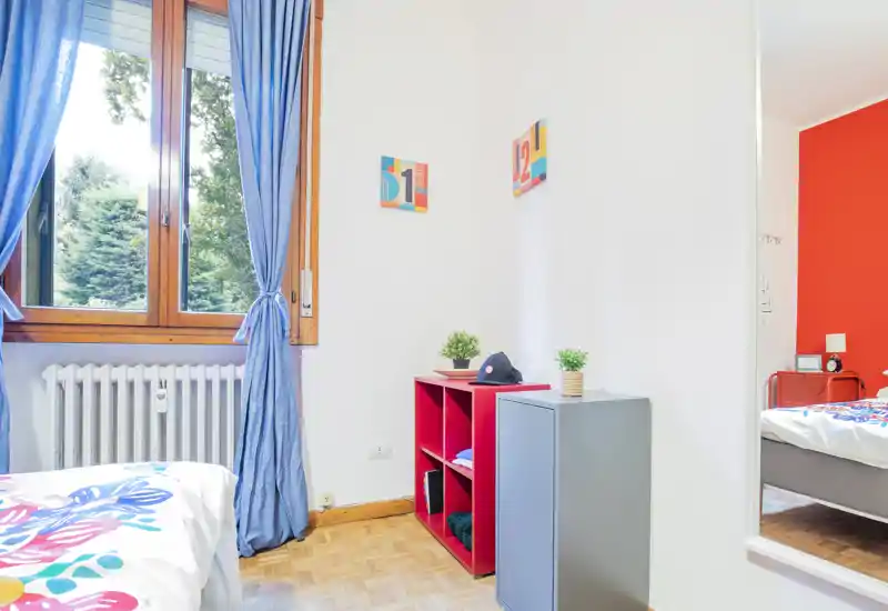 Bright, colorful bedroom with a comfortable bed visible at the left, a large window dressed with blue curtains, simple shelving and decorative plants — a well-lit, welcoming space ideal for a rental listing.