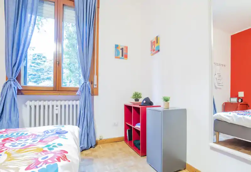Bright, colorful bedroom with a large window dressed in blue curtains, a partially visible bed with patterned bedding, and tidy storage shelves — ideal for a cheerful single or guest room.