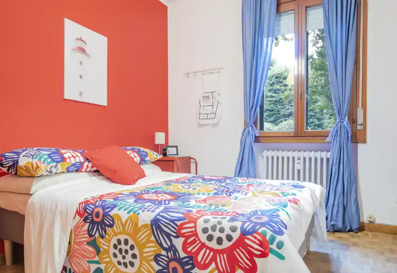 Bright, colorful bedroom with a comfortable double bed dressed in vibrant floral bedding, a red accent wall and natural light from a large window — stylish and inviting for short or long stays.