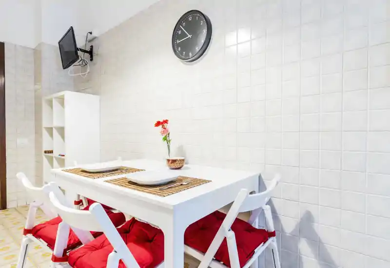 Bright, tidy dining corner with a white table set for two. Red cushions and simple decorative details create a welcoming, modern atmosphere.