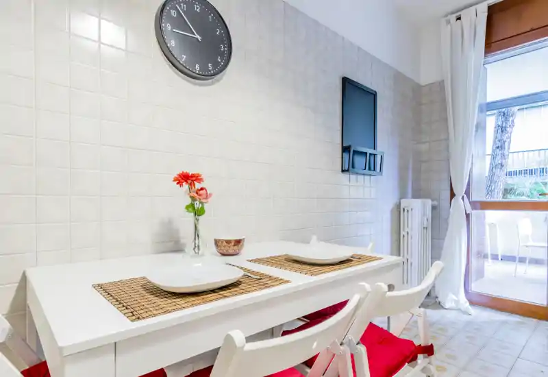 Bright, modern dining nook with a white table set for two, decorative placemats and a vase of flowers beside a large window leading to a balcony.