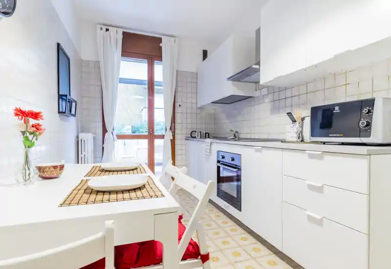 Bright modern kitchen with white cabinetry, built-in oven, microwave and a small dining table set beside a window leading to a balcony. Clean lines, decorative placemats and fresh flowers create a welcoming atmosphere ideal for short-term rentals.