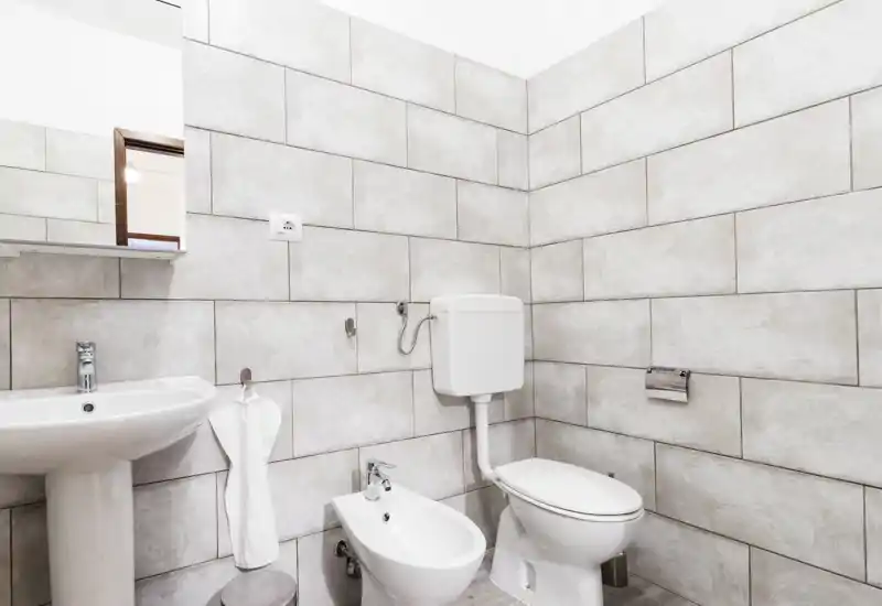 Bright, modern bathroom with wall-mounted cistern, bidet, toilet and pedestal sink against light stone tiles. Clean, minimal styling suitable for rental listings.