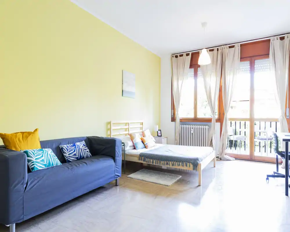 Dormitorio soleado y bien decorado con cama de madera, cojines decorativos y un sofá. Grandes ventanas con cortinas translúcidas inundan la habitación de luz natural, creando un ambiente acogedor.