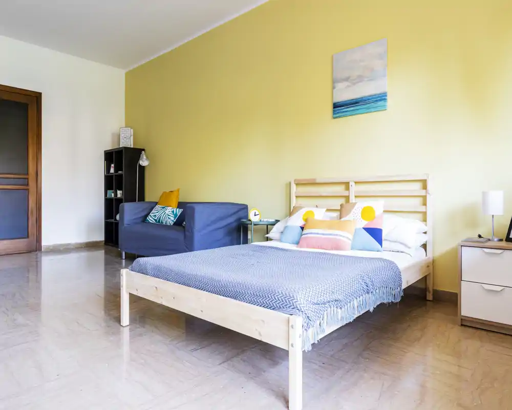 Dormitorio luminoso y moderno con cama doble de madera, cojines coloridos y manta azul; un pequeño sofá y mesitas de noche completan la distribución espaciosa.