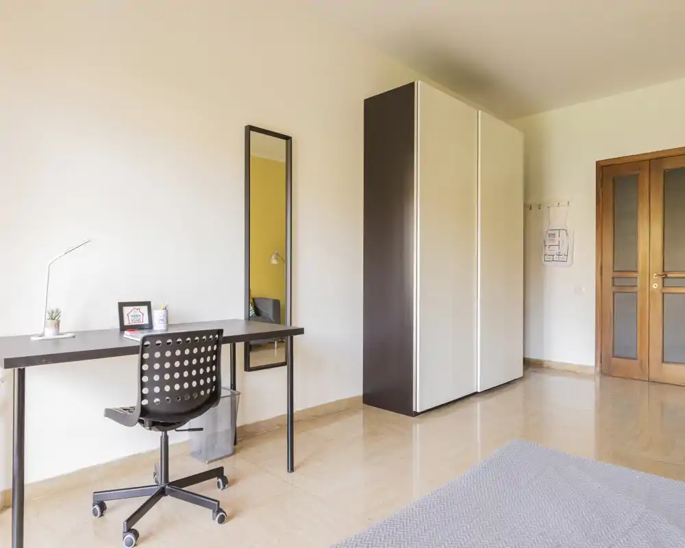 Rincón de dormitorio minimalista y luminoso con un escritorio, silla y armario; se aprecia una porción de la cama en primer plano. Tonos neutros y mobiliario moderno y sencillo crean un espacio funcional adecuado para alquileres temporales.