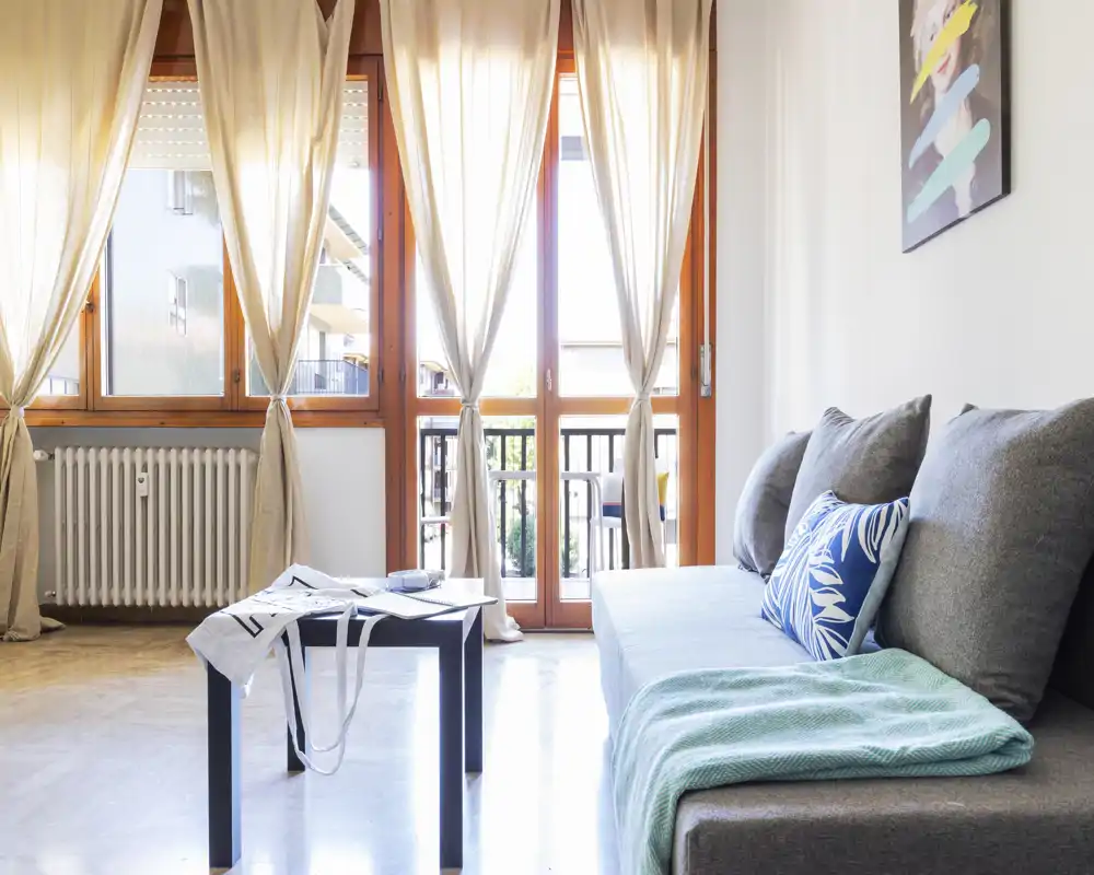 Soggiorno luminoso e moderno con divano grigio, cuscini decorativi e un piccolo tavolino davanti a grandi finestre che conducono a un balcone.