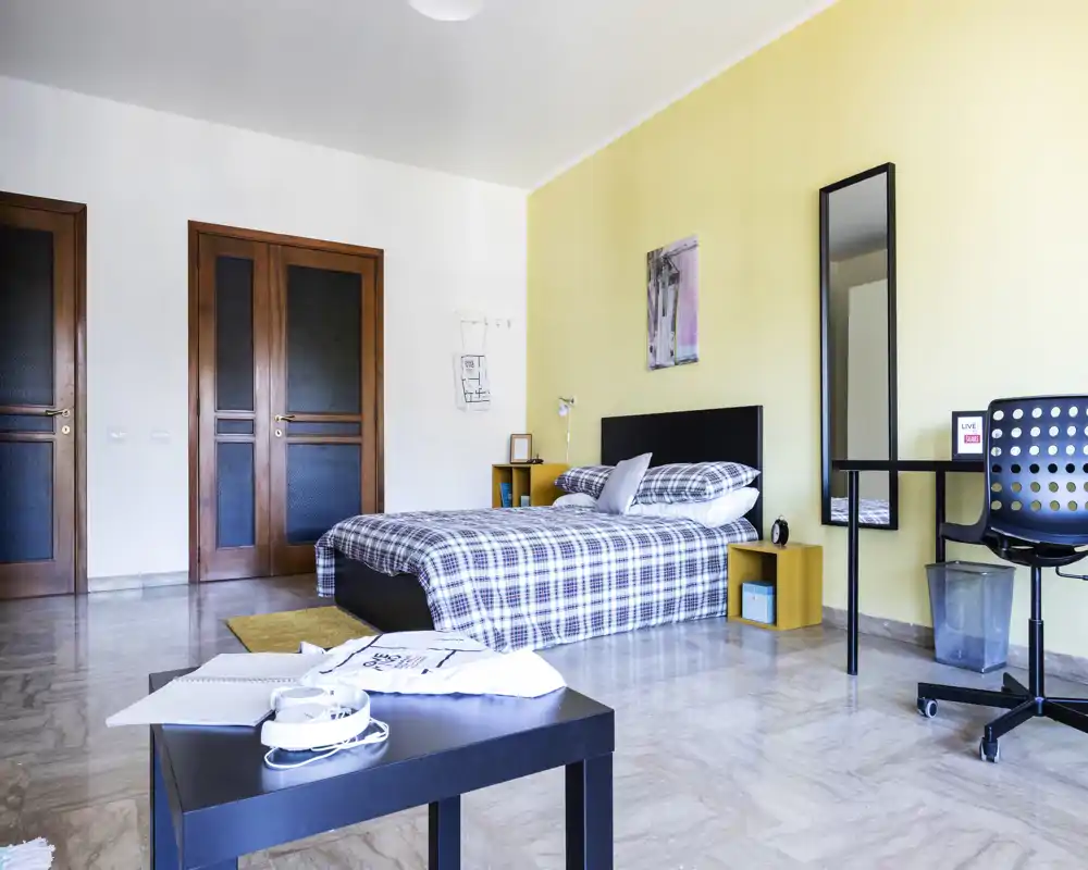 Camera da letto moderna e luminosa con letto matrimoniale con copriletto a quadri, comodini, specchio a figura intera e una scrivania. Parete gialla tenue, pavimento lucido e arredo minimale per un ambiente pulito e arioso.