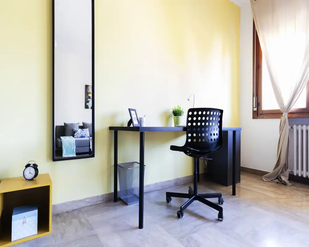 Zona giorno luminosa e moderna con scrivania minimal e sedia girevole forata accanto a una finestra soleggiata; dettagli curati come la piantina, lo specchio che riflette il divano con cuscini e il pavimento in marmo.