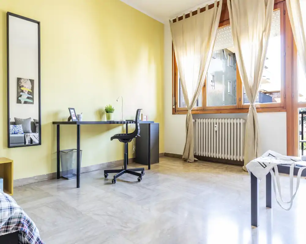 Camera da letto luminosa e minimale con scrivania vicino alla finestra, tende chiare e pavimento in marmo. In primo piano si vede parzialmente il letto; lo spazio è ben illuminato e ordinato, ideale per mostrare dimensioni e zona studio.