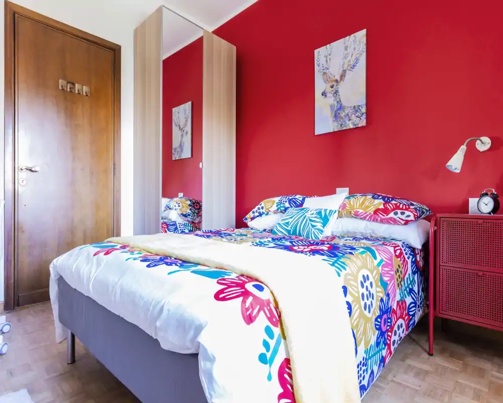 Dormitorio luminoso y colorido con cama doble vestida con ropa de cama floral, armario con espejo y mesita con lámpara y despertador — perfecto para fotos de alquiler.