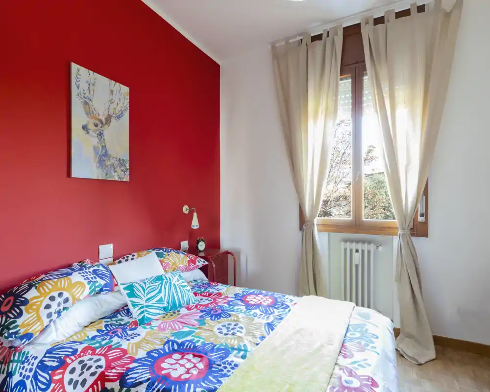 Dormitorio luminoso y colorido con cama doble frente a una pared roja, ropa de cama estampada y luz natural de una ventana grande.