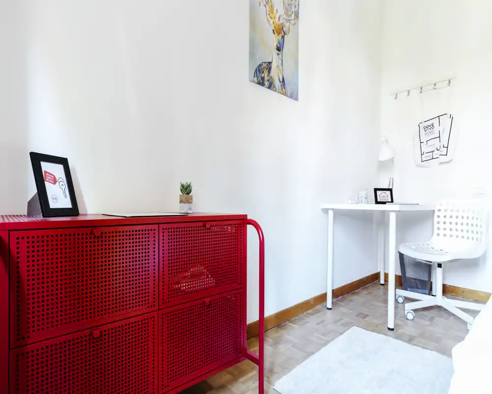 Rincón de dormitorio luminoso y minimalista con un llamativo armario rojo, pequeño escritorio blanco y silla, alfombra decorativa y cuadro: espacio limpio y moderno perfecto para un alquiler compacto.