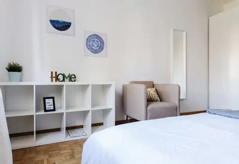 Bright, minimal bedroom with a neatly made bed in the foreground, a cozy armchair and open cubed shelving — calm white walls and wood flooring create a fresh, inviting atmosphere.