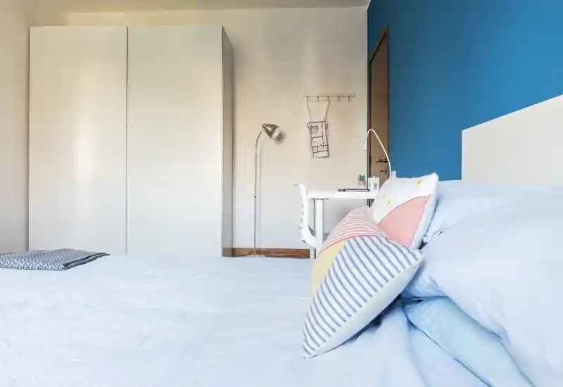 Bright, modern bedroom with a light-blue bed, colorful cushions and a white wardrobe; a small desk and floor lamp sit against a bold blue accent wall.