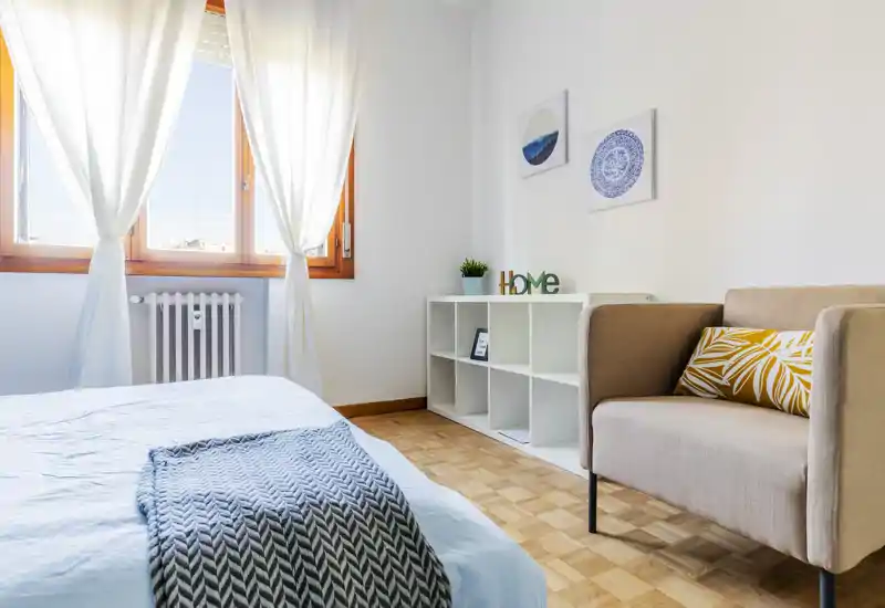 Bright, modern bedroom with natural light streaming through a large window, a neatly made bed with a textured throw, cozy armchair and simple shelving — ideal for rental photos.
