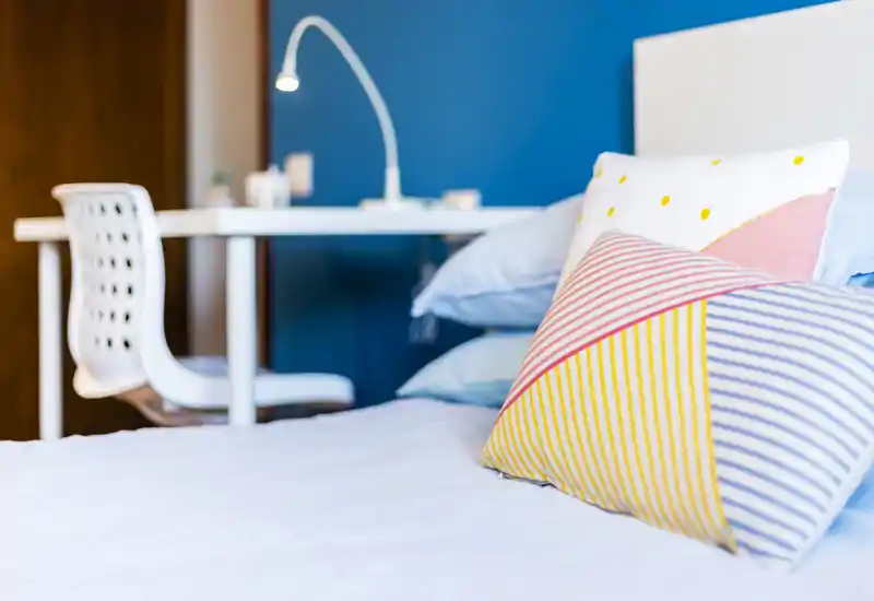 Bright, styled bedroom shot showcasing colorful cushions on a neatly made bed with a study desk and lamp in the soft-focused background.