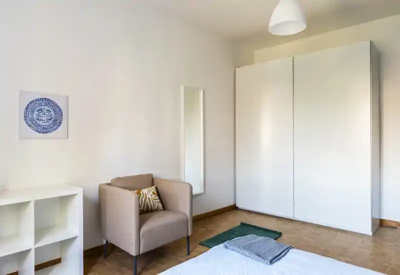 Bright, minimal bedroom with a visible bed corner, wardrobe and a cozy armchair; neutral tones and simple decor create a calm atmosphere ideal for rental photos.