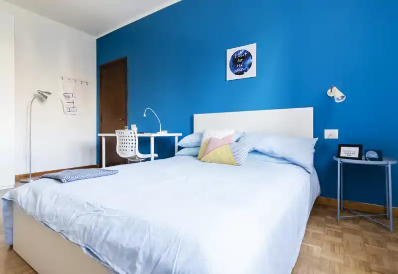 Bright, modern bedroom with a bold blue accent wall, neatly made double bed and a small workspace. Natural light and simple, colorful cushions create an inviting atmosphere ideal for short-term rentals.