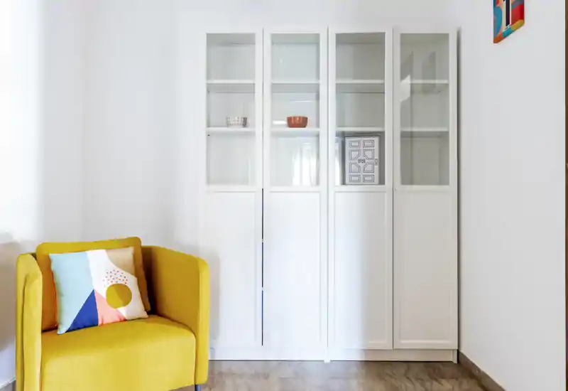 Bright, modern living area with a vibrant yellow armchair, decorative cushion and a tall white display cabinet — ideal for showcasing a cozy corner in a rental listing.