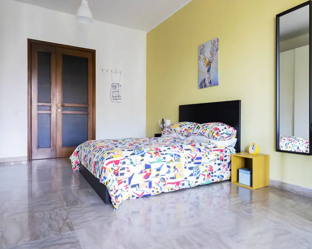Camera da letto luminosa e moderna con copriletto colorato a fantasia, letto matrimoniale basso, comodino e grande specchio a parete. Pavimento in pietra levigata, porta in legno e parete accentata gialla.