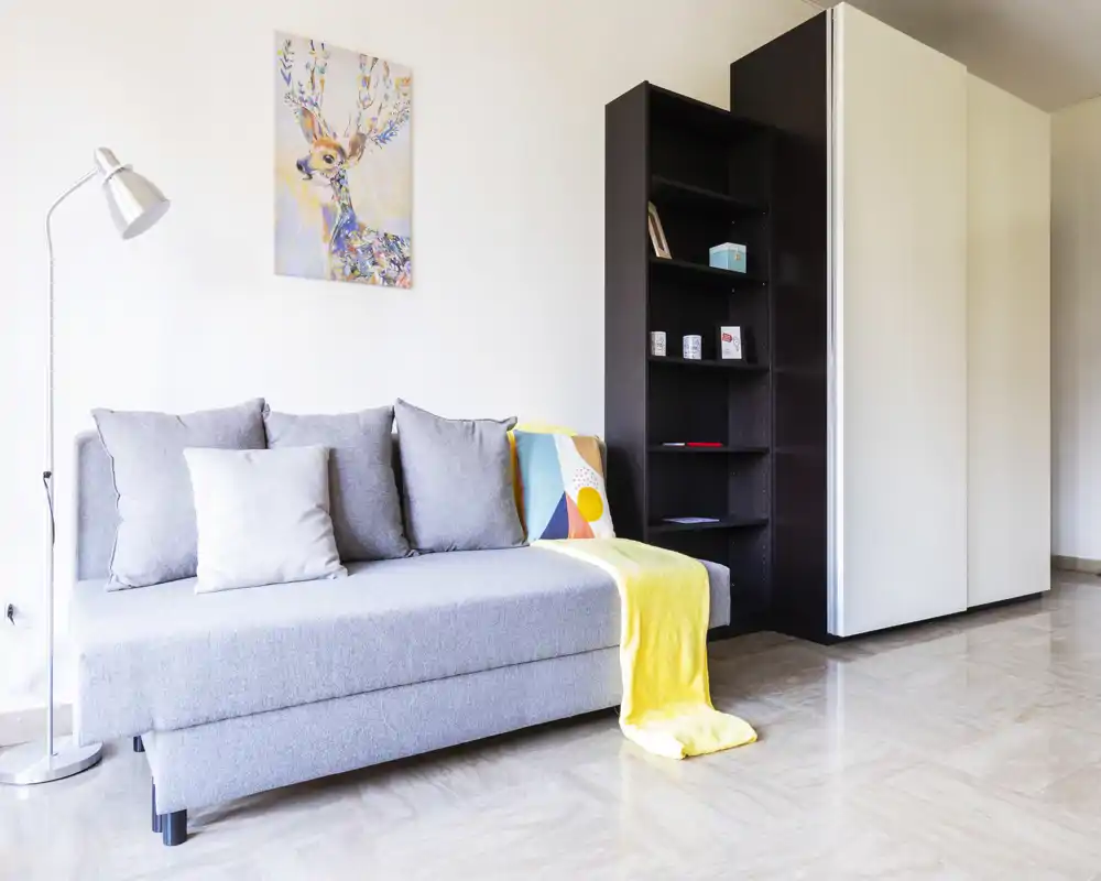 Zona giorno moderna e luminosa con divano grigio chiaro, cuscini decorativi, plaid colorato e scaffalatura—arredamento pulito e accogliente ideale per affitti.