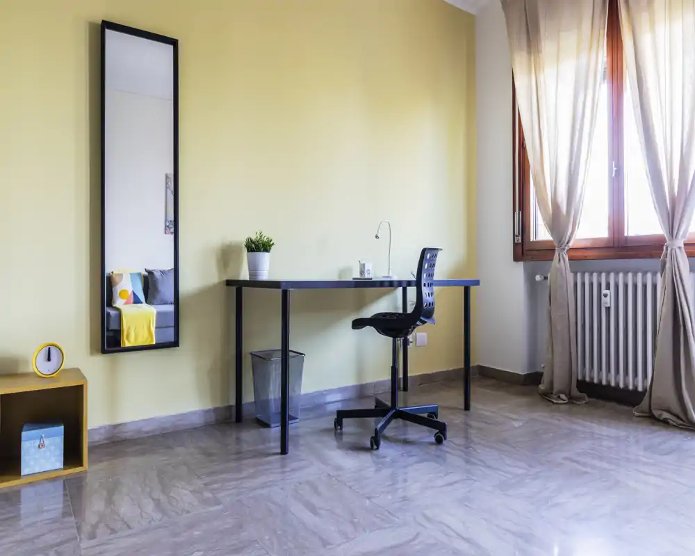 Zona giorno luminosa e minimal con una scrivania sottile, sedia girevole e una grande finestra che lascia entrare luce naturale; uno specchio verticale e piccoli dettagli decorativi donano un aspetto moderno e arioso.