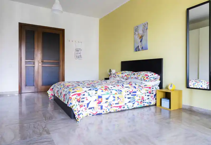 Camera da letto luminosa e moderna con copriletto colorato a fantasia, letto matrimoniale basso, comodino e grande specchio a parete. Pavimento in pietra levigata, porta in legno e parete accentata gialla.